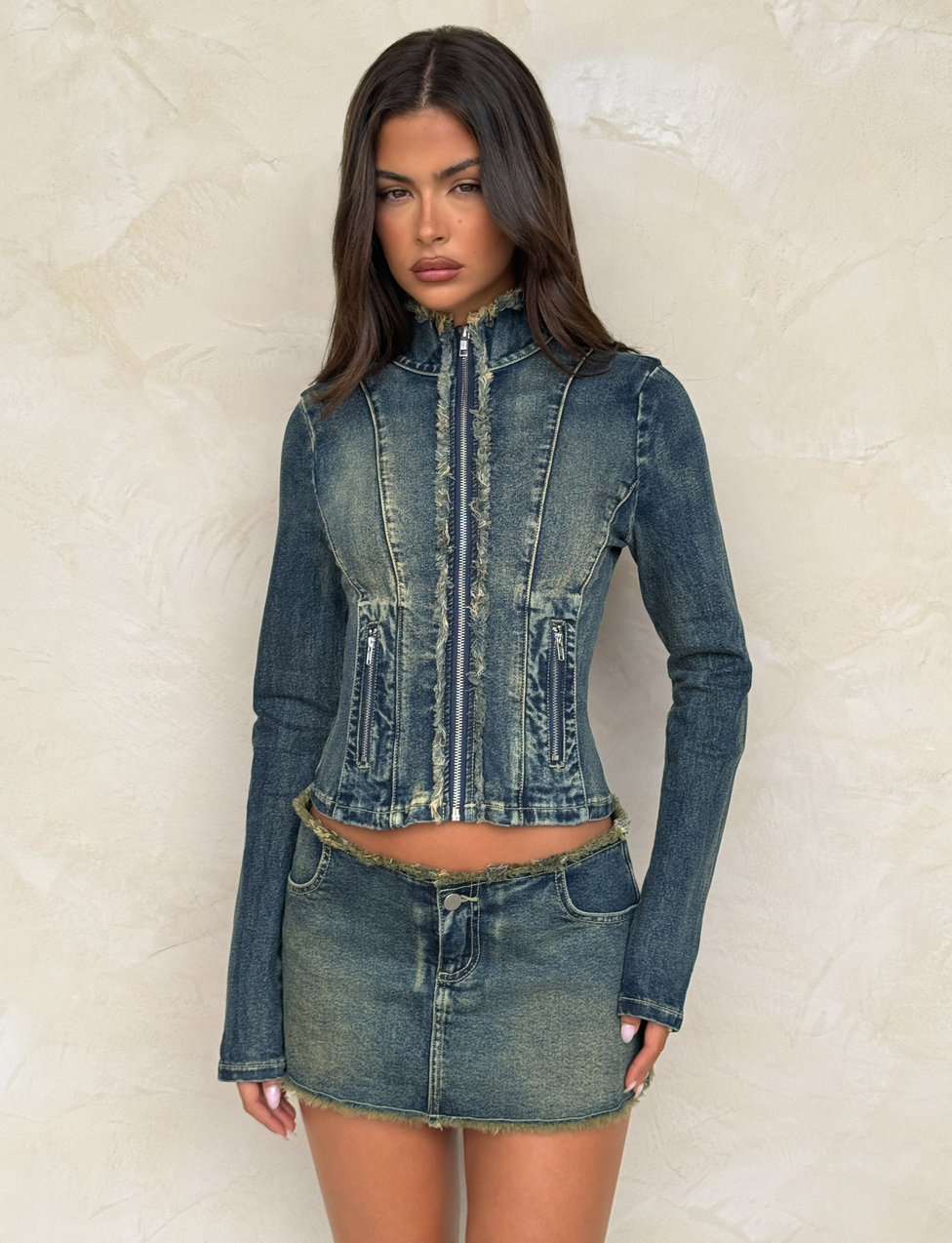 A woman poses against a beige textured wall, wearing the TIGERMIST OPHELIE JACKET in denim blue with frayed edges, paired with a matching short denim skirt.