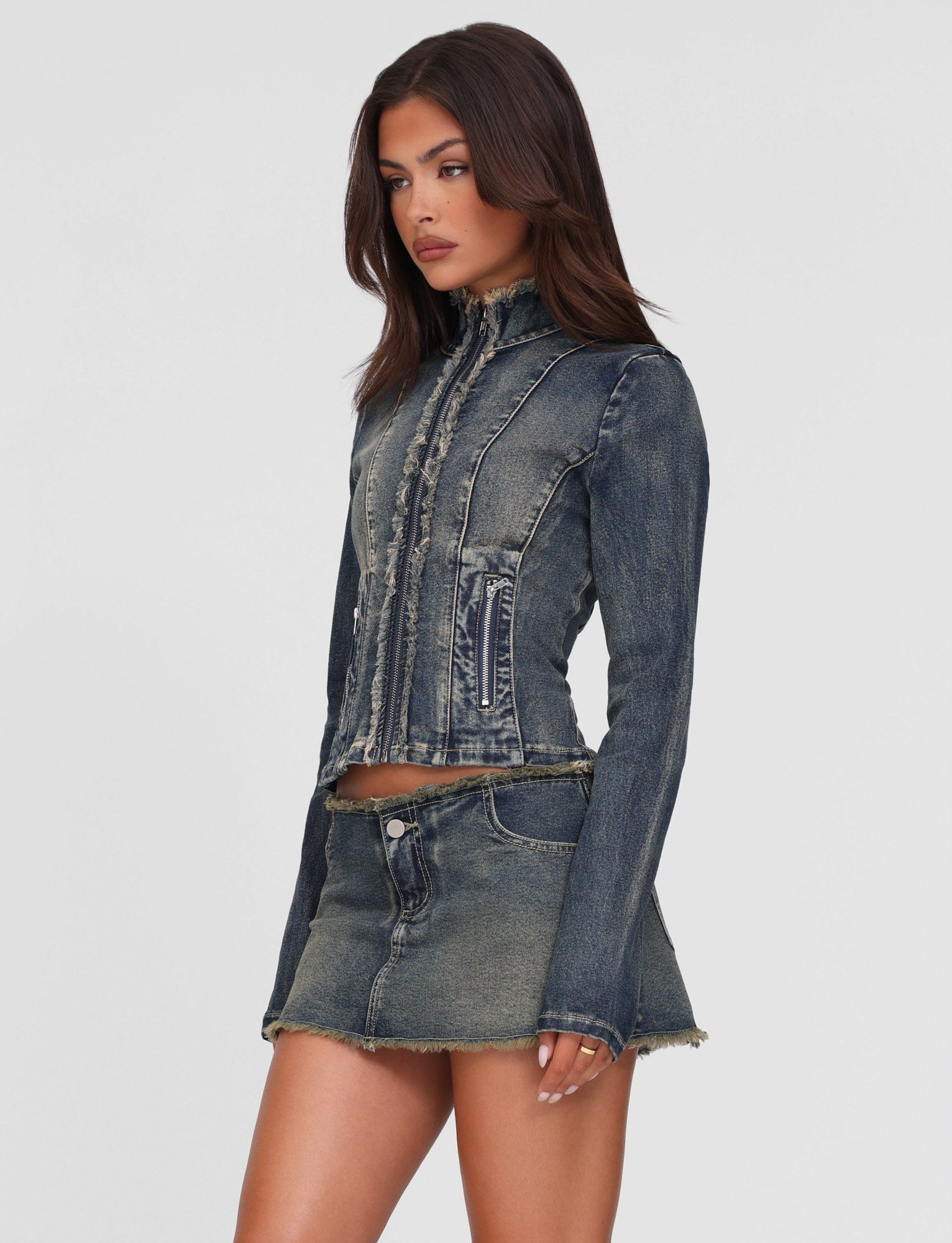A woman with long brown hair and a neutral expression models the TIGERMIST OPHELIE JACKET in denim blue, featuring a vintage wash and frayed edges, paired with a matching mini skirt against a white background.