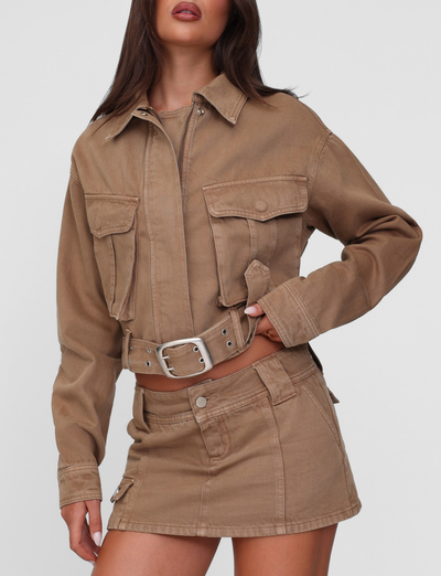 A woman wears the TIGERMIST ZELIE JACKET in washed tan, a neutral cropped utility style with patch pockets, paired with a matching high-waisted mini skirt featuring a silver buckle belt, set against a plain light background.