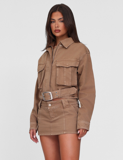 A woman with long dark hair and a neutral expression models the TIGERMIST ZELIE JACKET - NEUTRAL : SANDBLAST : WASHED TAN, featuring functional patch pockets, paired with a matching mini skirt against a plain white background.