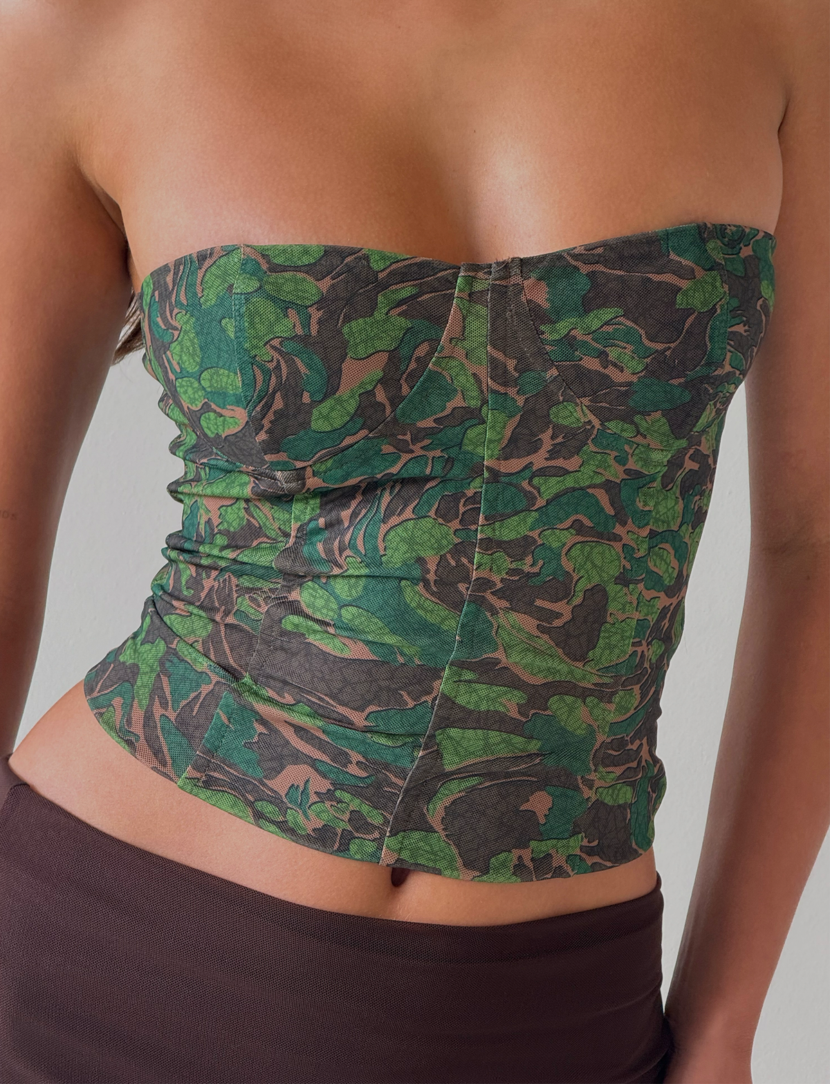 A person wearing the TIGERMIST DULCE CORSET - GREEN : CAMO, a strapless fitted camo top with shaping effect, paired with a brown skirt and standing against a plain light background.