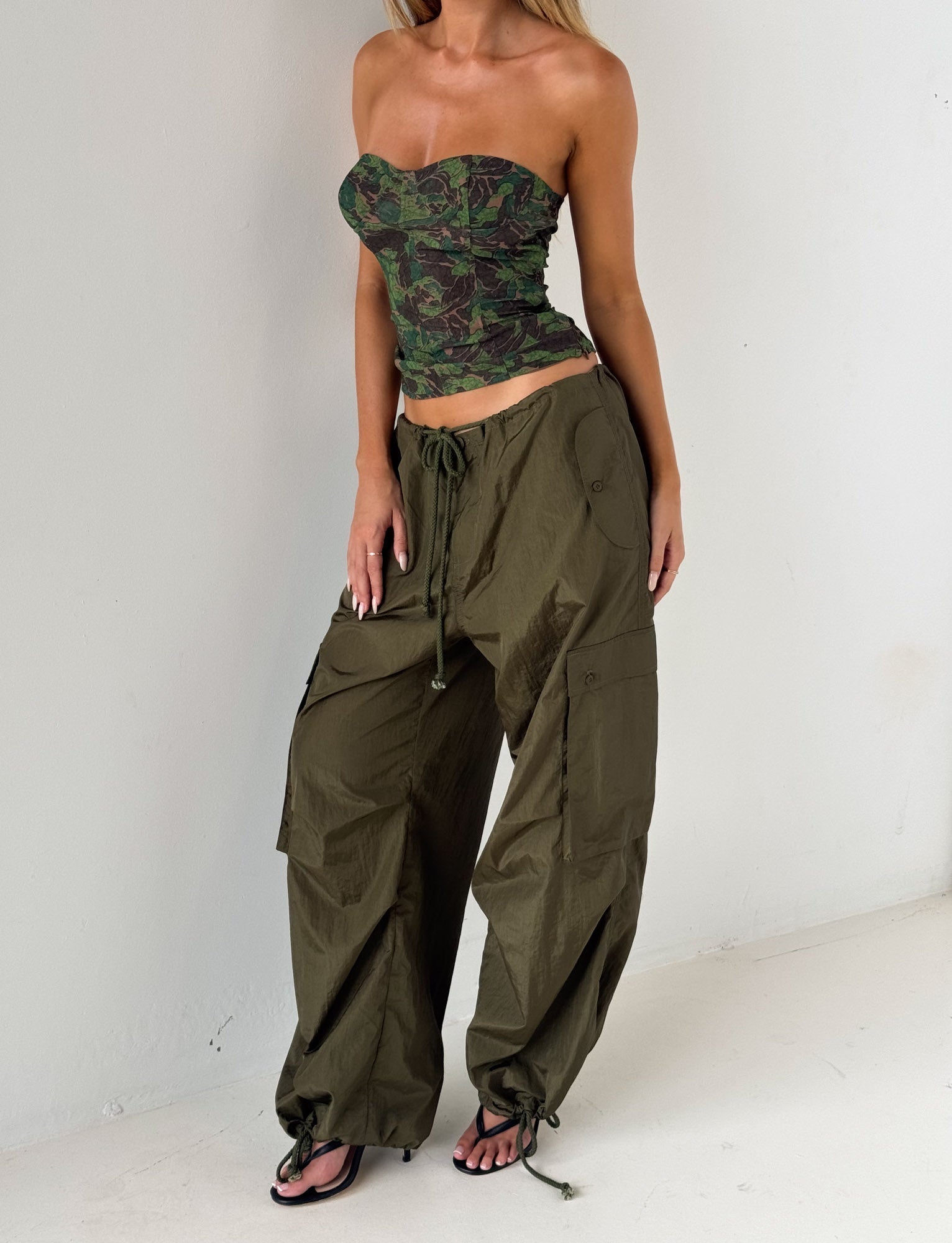 A woman wearing the TIGERMIST VIOLETTE PANT in khaki green stands against a white wall, pairing the loose olive cargo pants with a green camouflage strapless top and resting her hand on her hip.
