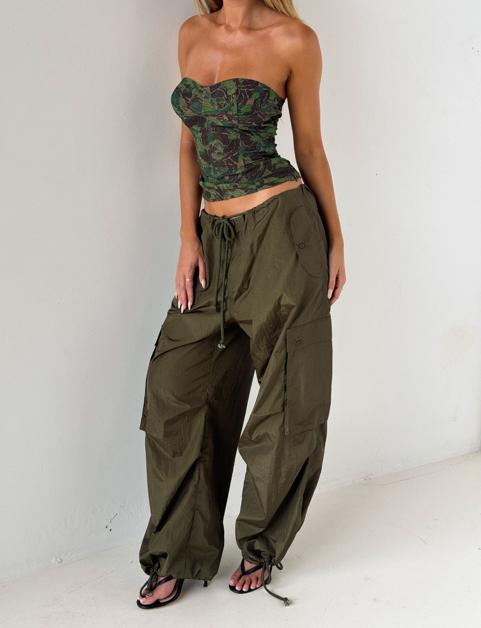 A woman wearing the TIGERMIST VIOLETTE PANT in khaki green stands against a white wall, pairing the loose olive cargo pants with a green camouflage strapless top and resting her hand on her hip.