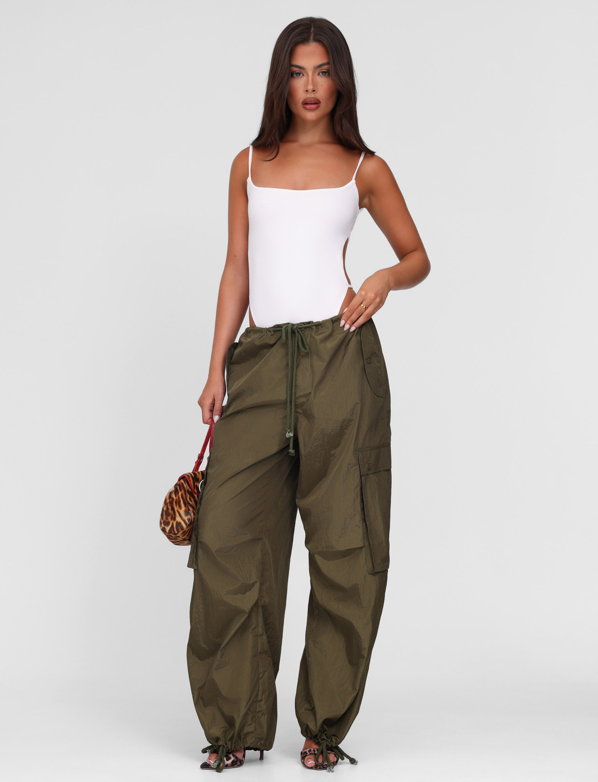 A woman with long brown hair and a neutral expression wears the TIGERMIST VIOLETTE PANT in khaki green, paired with a white sleeveless bodysuit and holds a small animal print handbag, standing against a plain background.