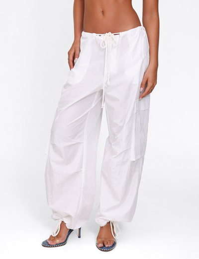 A person models TIGERMIST's VIOLETTE PANT in white—low rise, oversized fit, with drawstring waist—paired with blue high-heeled sandals, standing against a plain white background. The upper body is mostly out of frame.