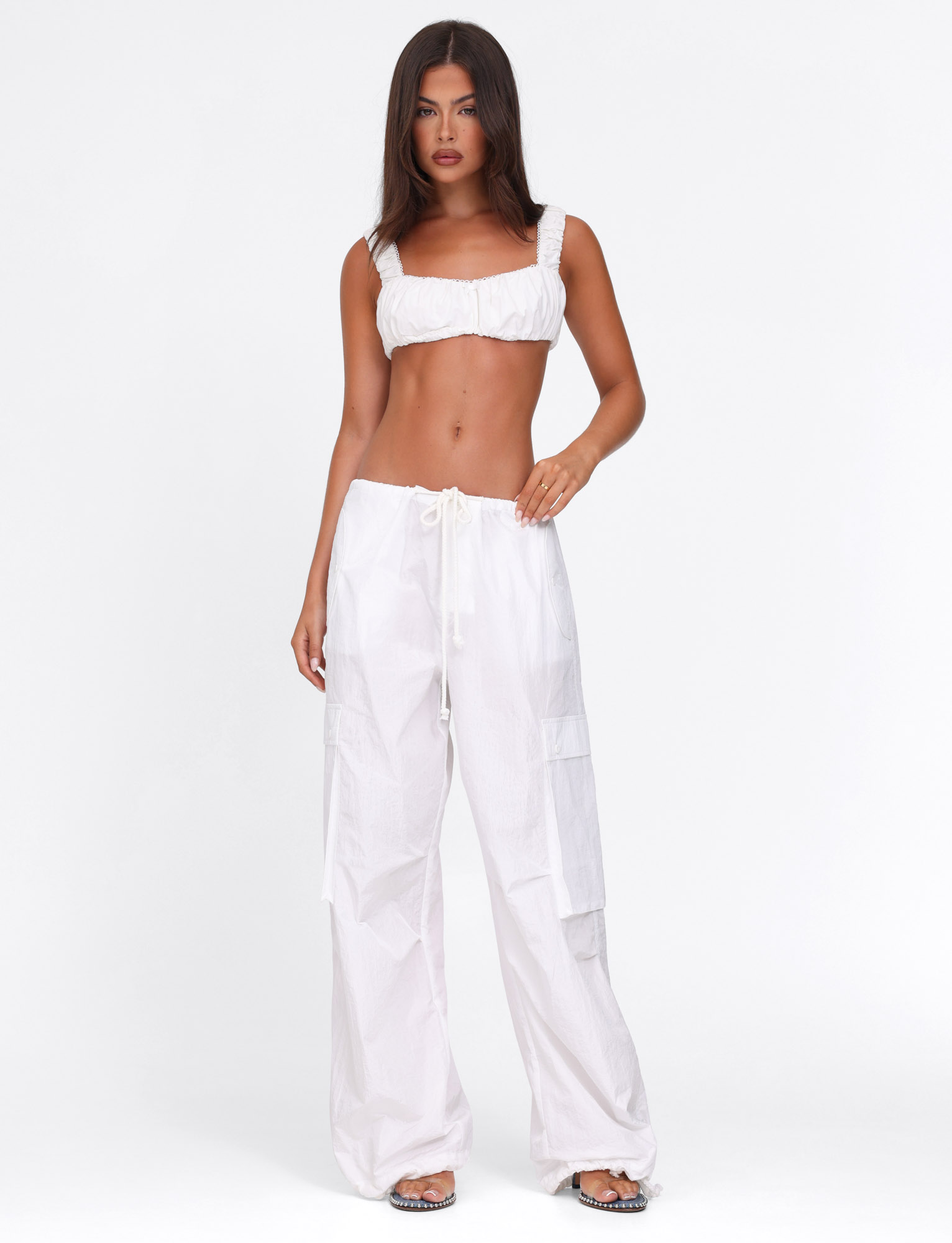 A woman with long, straight brown hair and a neutral expression stands against a white background wearing the TIGERMIST VIOLETTE PANT in white, styled with a white cropped top and flat sandals.