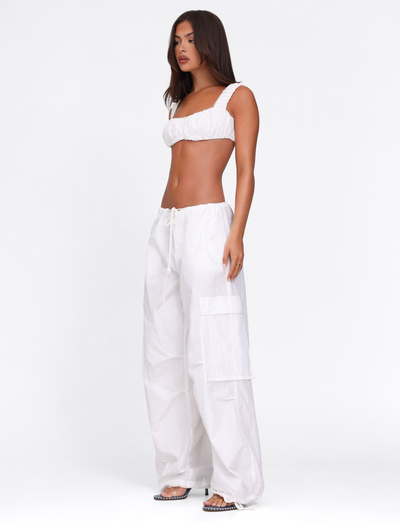 A woman with long straight brown hair and a neutral expression wears the TIGERMIST VIOLETTE PANT in white, styled with a white gathered-strap crop top and black open-toe sandals, standing against a plain white background.