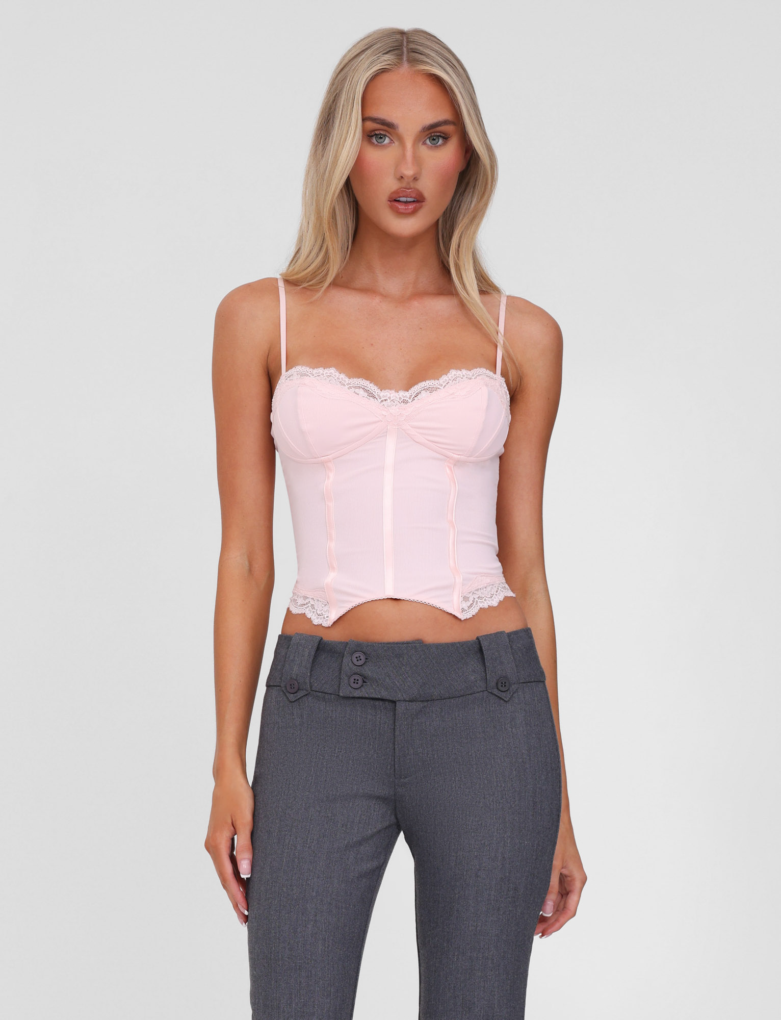A woman with long blonde hair wears the TIGERMIST NILA CORSET TOP in soft pink, featuring lace trim and adjustable straps, paired with high-waisted gray pants against a plain light background.