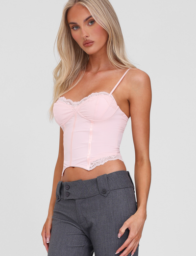 A woman with long blonde hair wears the TIGERMIST NILA CORSET TOP in soft pink, featuring lace trim and adjustable straps, paired with high-waisted gray trousers against a plain light gray background.