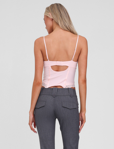 A woman with long blonde hair is seen from the back wearing the TIGERMIST NILA CORSET TOP in soft pink, paired with high-waisted gray pants featuring buttoned back pockets.