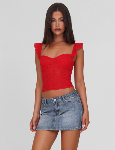 A woman with long dark hair models the TIGERMIST LORNA TOP in red, featuring frilled sleeves, paired with a short blue denim skirt against a plain light gray background.