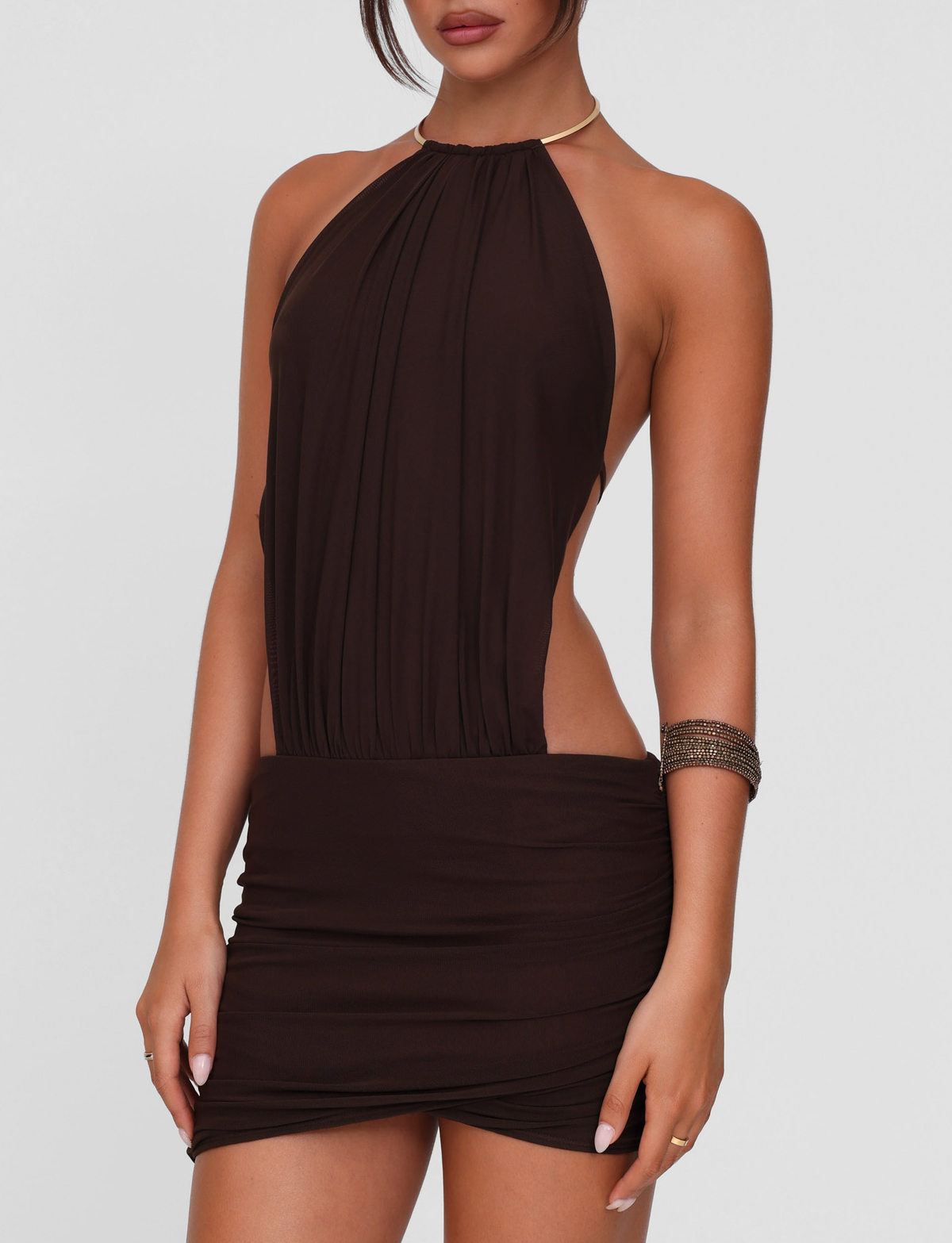 A woman models the TIGERMIST EVANNA MINI DRESS - BROWN : CHOCOLATE, a sleeveless, backless bodycon mini dress with waist ruching, styled with a gold necklace and metallic bracelet against a plain white background.