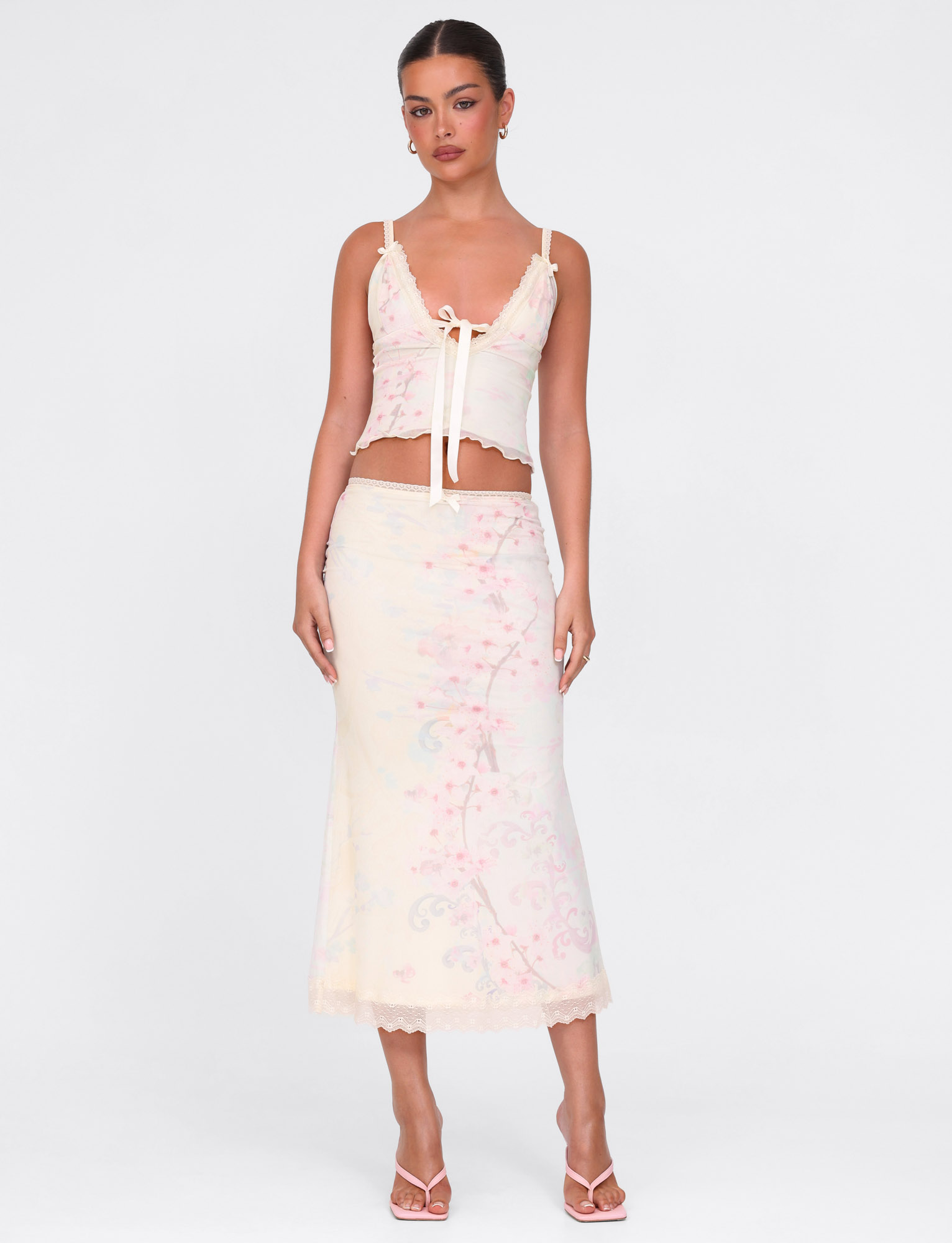 A woman stands against a white background wearing the TIGERMIST LONIE TOP in Neutral Floral Washed Cherry Blossom with a matching high-waisted skirt, paired with open-toe heels and simple earrings.