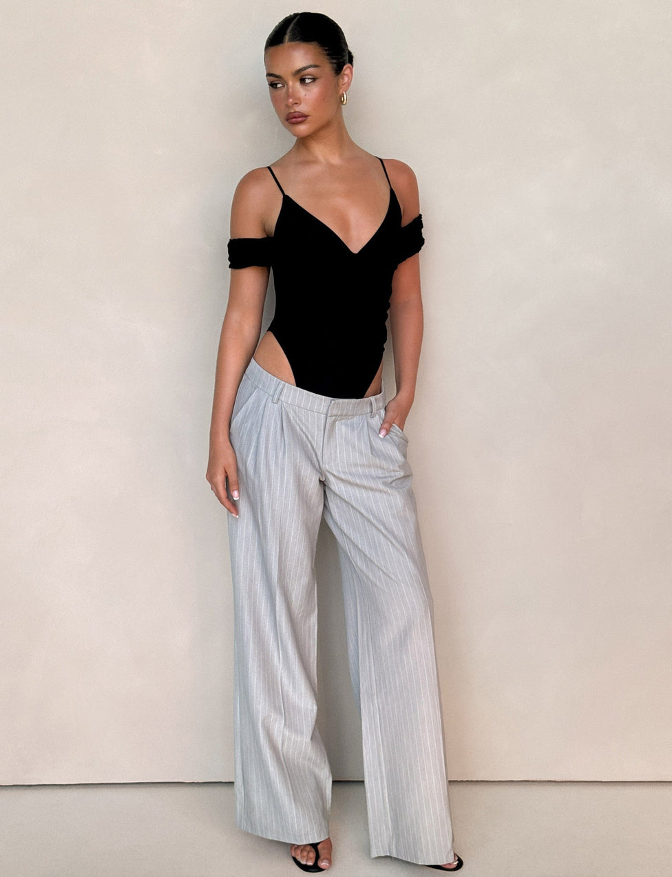 A woman stands against a neutral background in TIGERMIST’s AMBERLY SUIT PANT in grey pinstripe, paired with a black off-shoulder bodysuit and black heeled sandals. Her hair is up as she gazes slightly to the side.