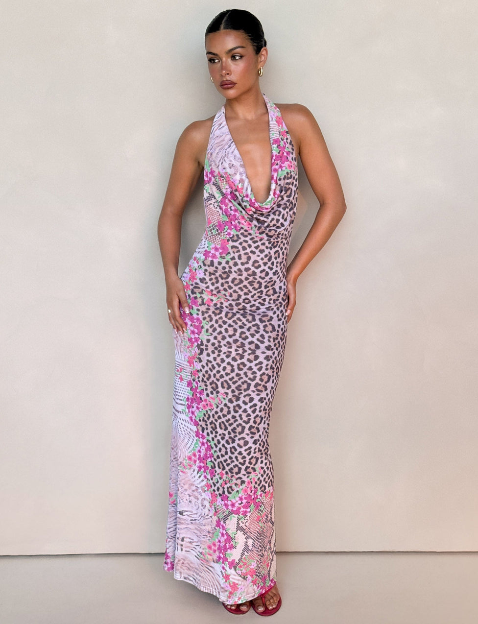 A woman stands against a beige wall wearing the TIGERMIST CATALINA MAXI DRESS in Neutral Animal Floral Border. The long, sleeveless dress features a pink, white, and gray floral leopard print. She pairs it with dark pink heels and has her hair pulled back.