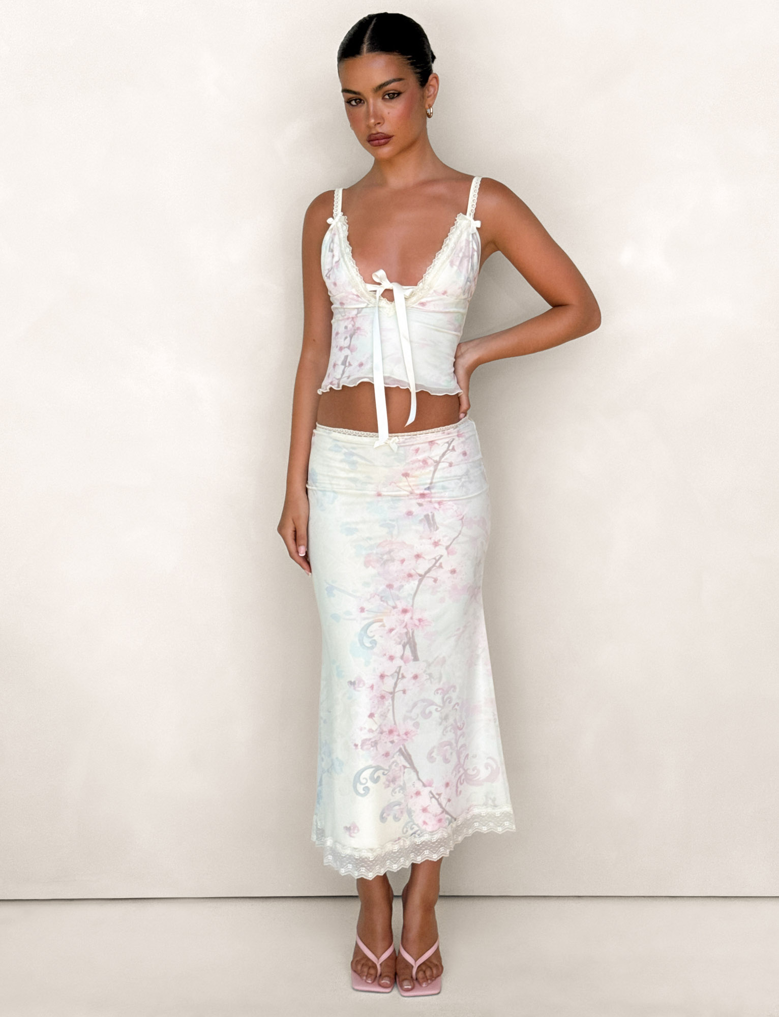 A woman stands against a plain background wearing the TIGERMIST LONIE MIDI SKIRT in Neutral Floral, Washed Cherry Blossom, paired with a matching fitted top featuring a bow accent. Her dark hair is pulled back and she wears pink heels.