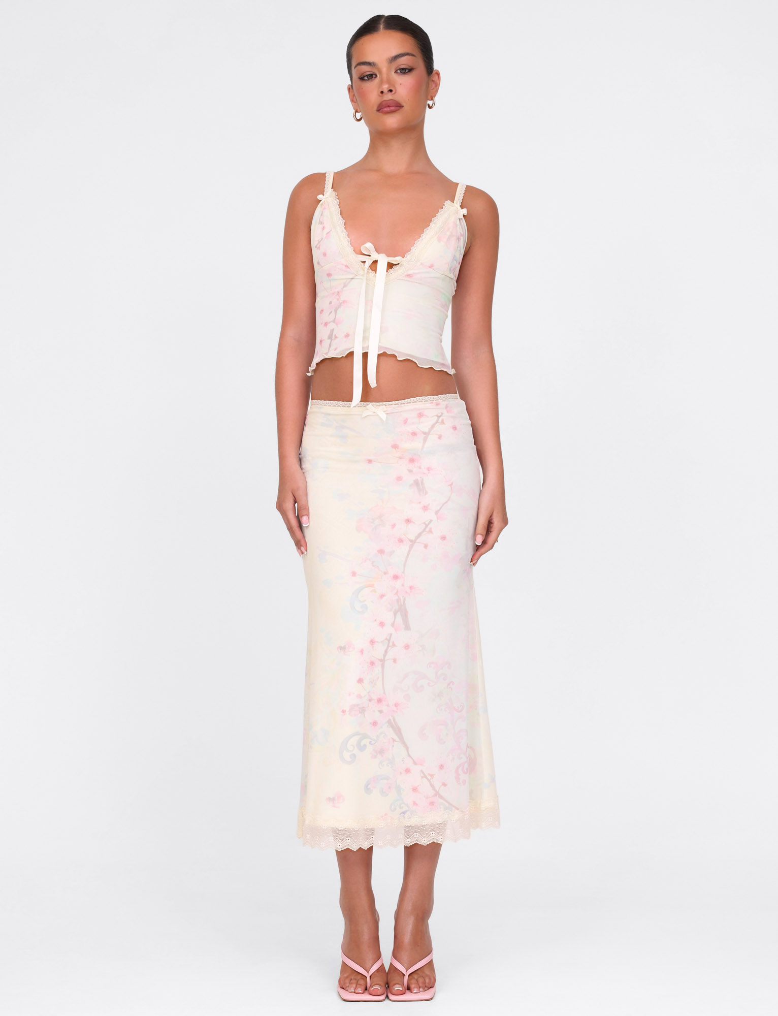A woman stands against a white background wearing the TIGERMIST LONIE MIDI SKIRT in Neutral Floral Washed Cherry Blossom, paired with a lace-trimmed tank top, heeled sandals, and her hair pulled back.