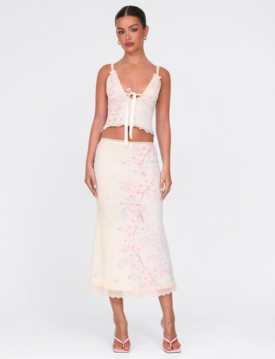 A woman stands against a white background wearing the TIGERMIST LONIE MIDI SKIRT in Washed Cherry Blossom and a matching pastel floral sleeveless tie-front top, with her hair tied back, earrings, and strappy heels.