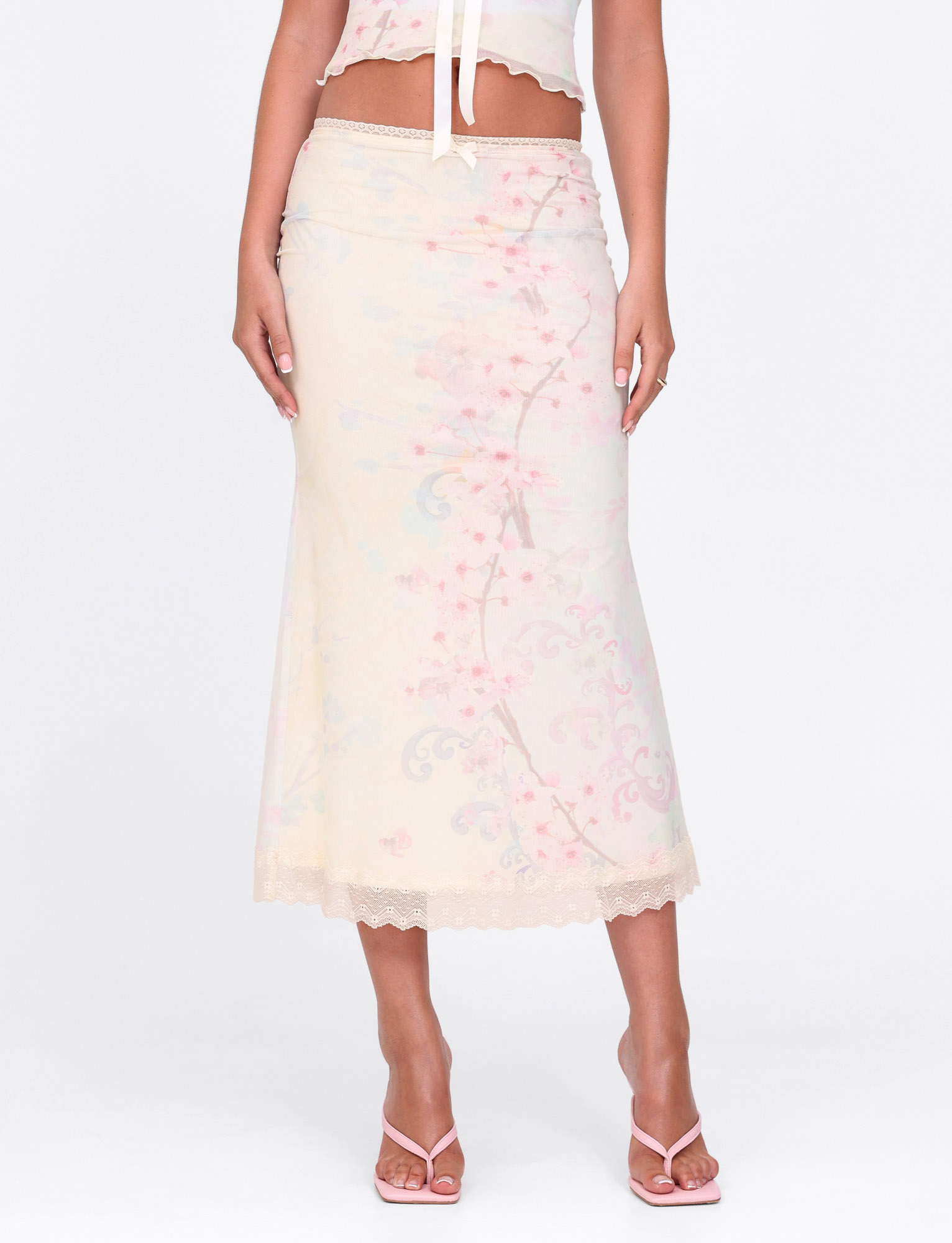 Wearing the TIGERMIST LONIE MIDI SKIRT in Washed Cherry Blossom floral, paired with a matching cropped top and white heeled sandals, a person stands against a plain white background.