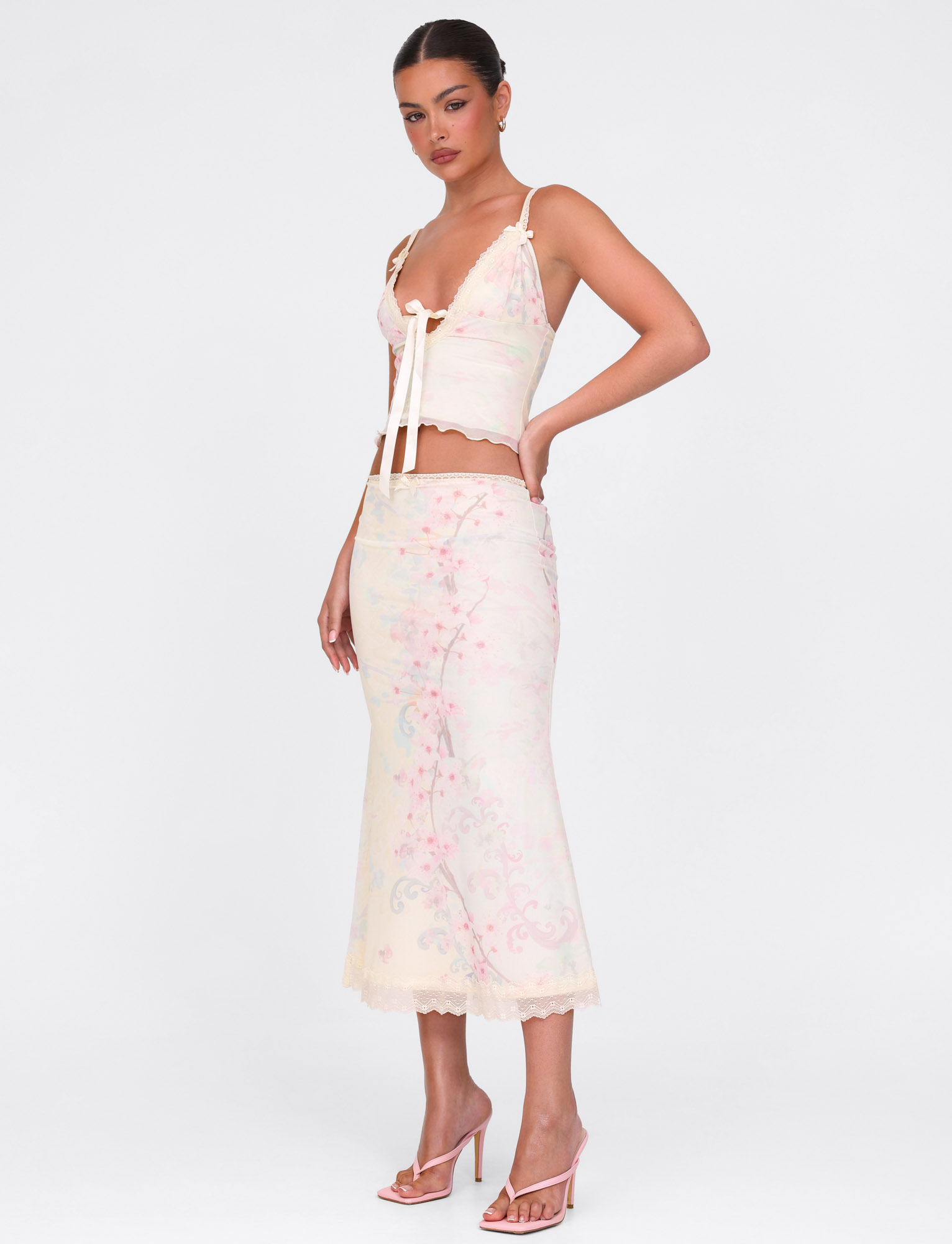 A woman stands against a white background in a pastel floral crop top and the TIGERMIST LONIE MIDI SKIRT in Neutral Floral Washed Cherry Blossom, her hair tied back, finished with pink high-heeled sandals.