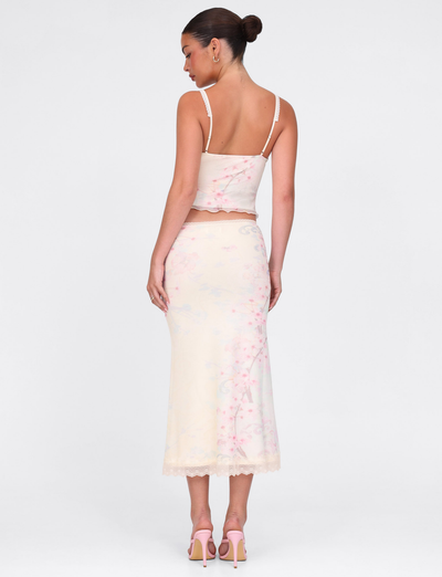 A woman with her hair in a bun faces away, wearing the TIGERMIST LONIE MIDI SKIRT in Neutral Floral Washed Cherry Blossom, paired with a matching lace-trim camisole and pink heels against a plain white background.
