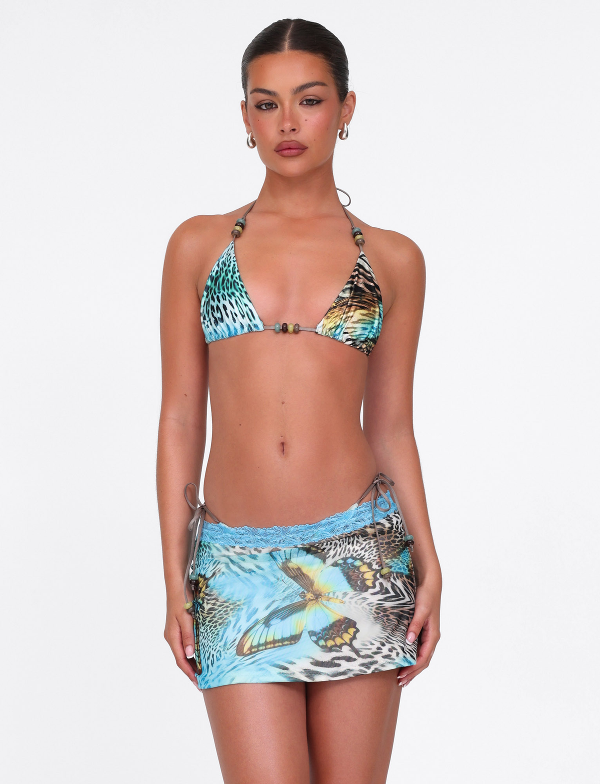 A woman in the TIGERMIST FINLEA MINI SKIRT - MULTI : ANIMAL : BUTTERFLY ANIMAL and a matching butterfly animal print bikini top with beaded straps stands against a white background, looking at the camera with a neutral expression.