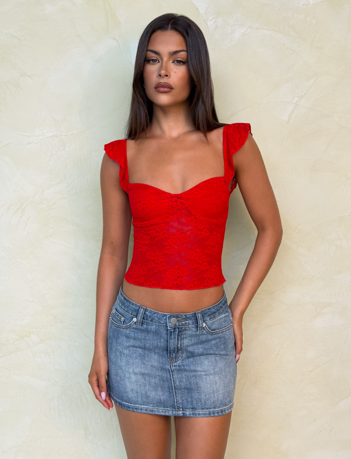 A woman with long, straight dark hair and a neutral expression stands against a light textured wall, wearing a red lace sleeveless top with ruffled straps and the TIGERMIST MICHAYLA MINI SKIRT - MID WASH.