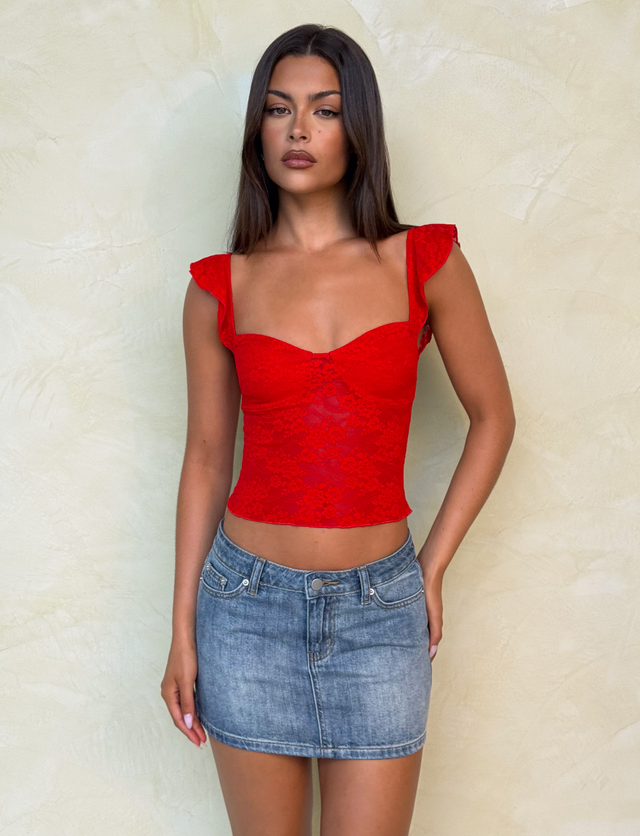 A woman with long, straight dark hair and a neutral expression stands against a light textured wall, wearing a red lace sleeveless top with ruffled straps and the TIGERMIST MICHAYLA MINI SKIRT - MID WASH.