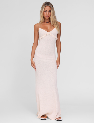 A woman poses against a plain background in the TIGERMIST LAVELLE MAXI DRESS in cream, featuring thin crisscross straps and a floral chest accent. She has long, loose blonde hair and neutral makeup.