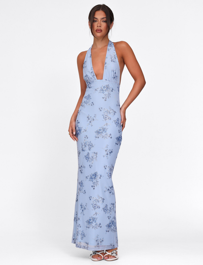 A woman models the TIGERMIST PHAEDRA MAXI DRESS in blue floral, featuring a sleeveless design, deep V-neckline, and metallic foil detail, standing against a plain white background.