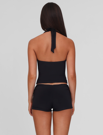 A woman with long brown hair stands with her back to the camera, wearing a black halter top and TIGERMIST JOVIENNE SHORT - BLACK jersey mini shorts with a fitted silhouette, set against a plain light gray background.