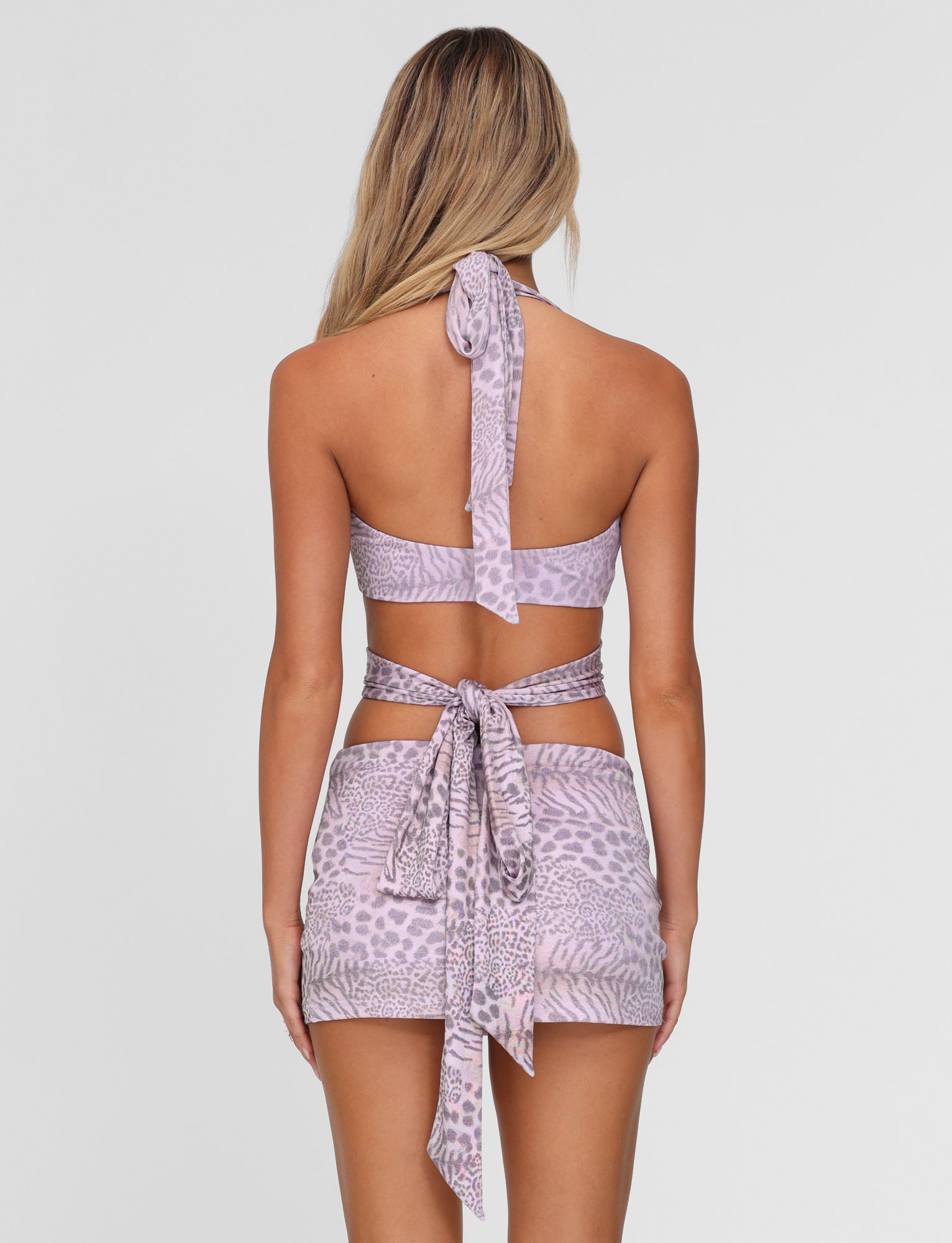 A woman with long, blonde hair is seen from behind wearing the TIGERMIST EVANGELINE TOP in pale pink tiger print, tied at the neck and back, paired with a matching bodycon mini skirt cinched at the waist.