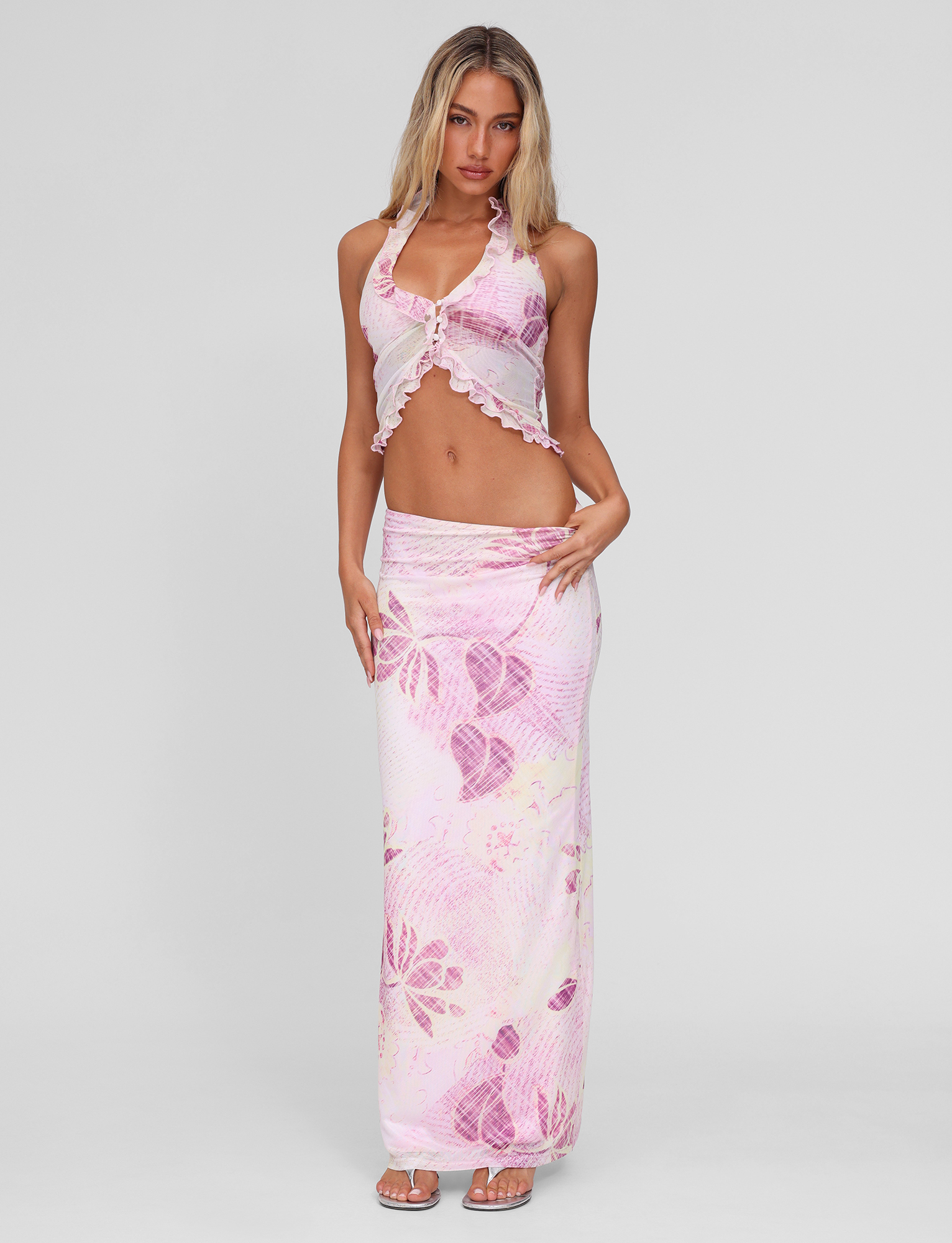 A woman stands against a plain background wearing the TIGERMIST DALILA MIDI SKIRT in Grunge Floral Magenta with a matching sleeveless halter crop top, both featuring ruffle trim, paired with sandals.