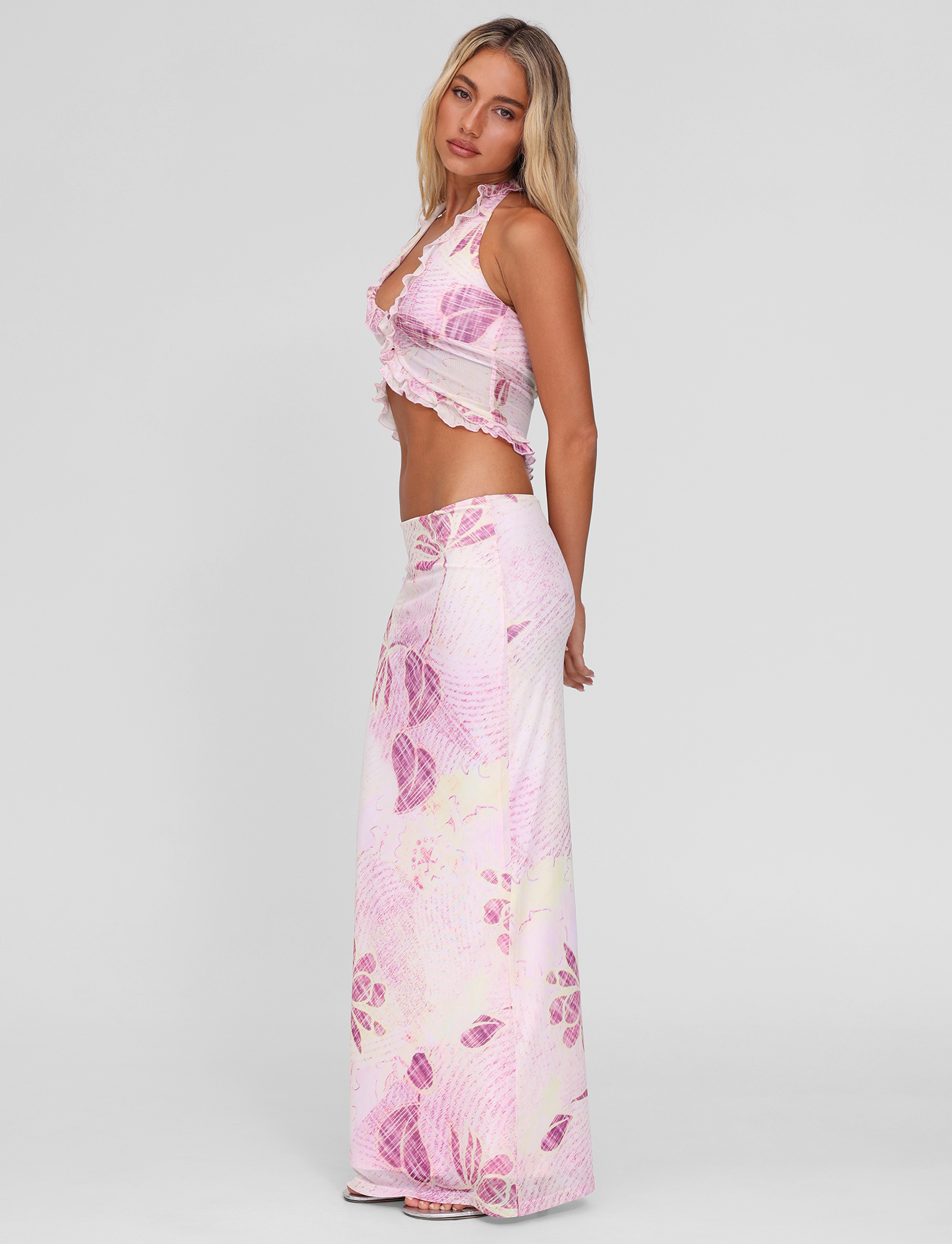 A woman with long blonde hair stands sideways against a plain background, wearing the TIGERMIST DALILA MIDI SKIRT in Grunge Floral Magenta with a matching sleeveless, ruffled crop top.