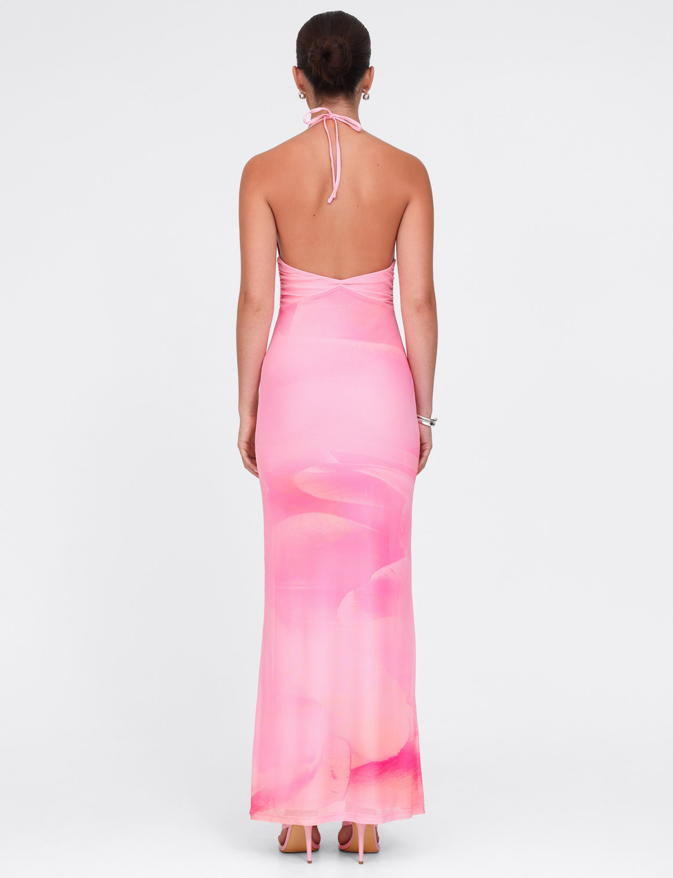 A woman faces away, wearing the TIGERMIST DENAE MAXI DRESS in Pink Blossom— a long, fitted pink lace halter dress with an open back. Her hair is up; she pairs it with bracelets and nude heels against a plain white background.