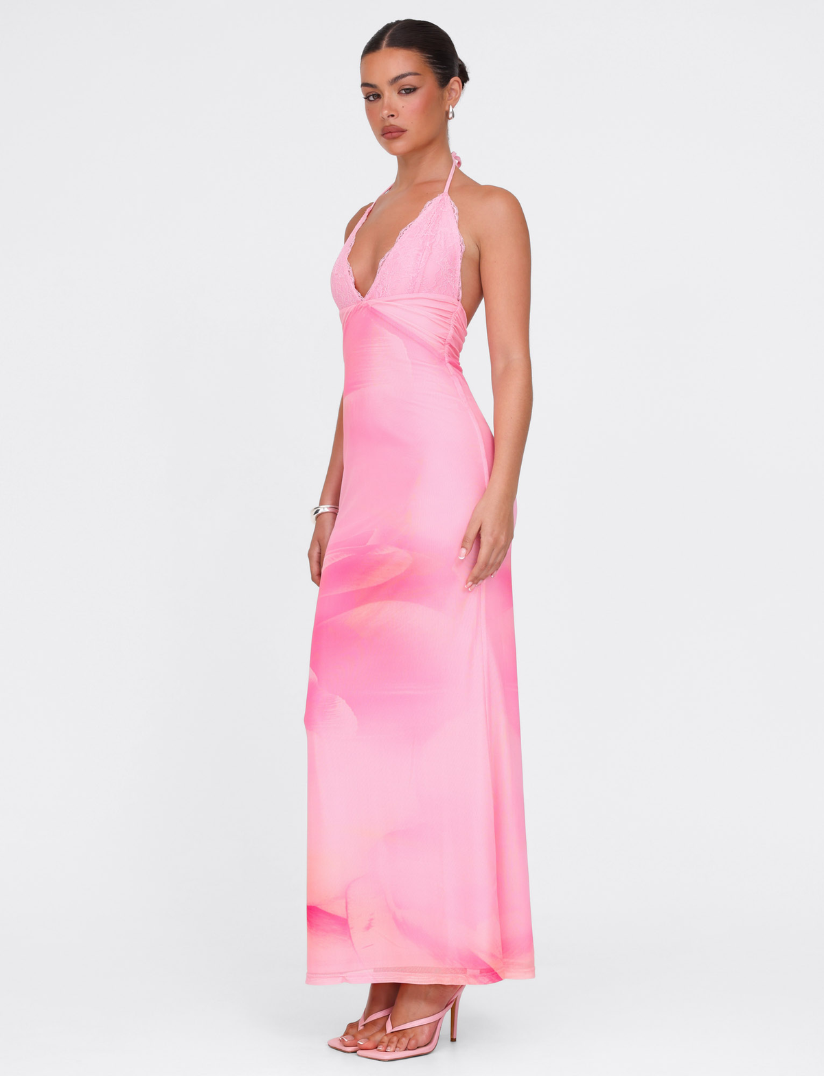 A woman stands in profile against a white background wearing the TIGERMIST DENAE MAXI DRESS in Pink Blossom—a floral pink lace halter dress with a deep V neckline—paired with pink high-heeled sandals.