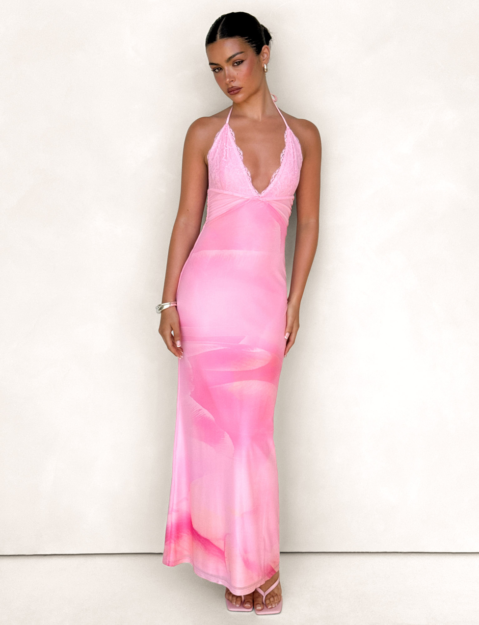 A woman poses confidently against a plain light background, wearing the TIGERMIST DENAE MAXI DRESS in Pink Blossom with strappy sandals, sleek hair, and subtle jewelry.