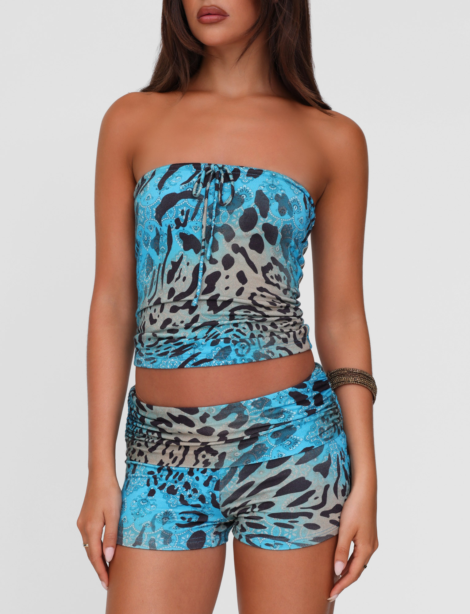 A woman wears the TIGERMIST MIRELY TOP - MULTI: LEOPARD SWIRL and matching shorts, both in a fitted blue leopard print. The high-stretch set flatters her figure as she stands against a white background, her face partly out of frame.