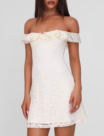A woman wears the TIGERMIST DENALI LACE MINI DRESS in cream lace, featuring off-shoulder style and ruffled neckline, fitted through the body, standing against a plain light background.