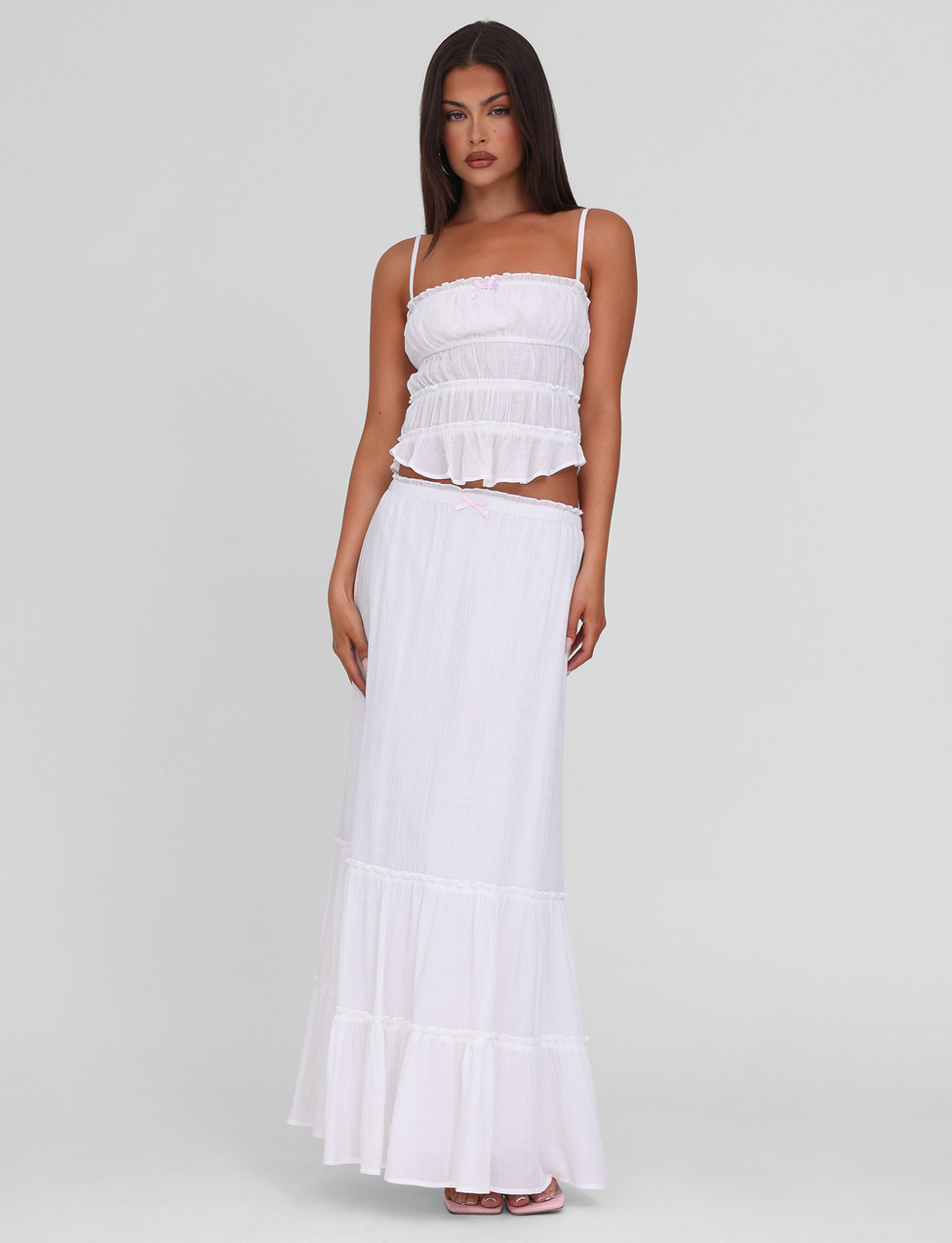 A woman with long dark hair and a neutral expression stands against a plain background wearing the TIGERMIST ASHLYNN TOP in white, featuring adjustable shoulder straps and tiered detailing. One hand rests by her side as she pairs it with a flowing white skirt.
