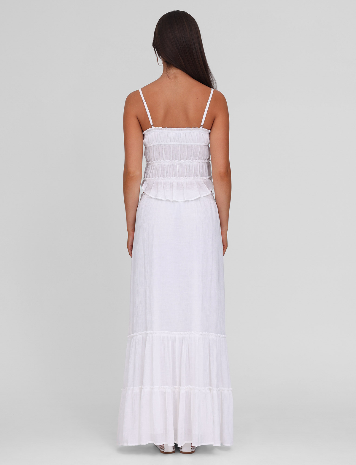 A woman with long dark hair, seen from behind, wears the TIGERMIST ASHLYNN MAXI SKIRT in white. The sleeveless skirt has thin straps, a ruffled tiered design, elasticated waist, and is set against a plain light gray background.