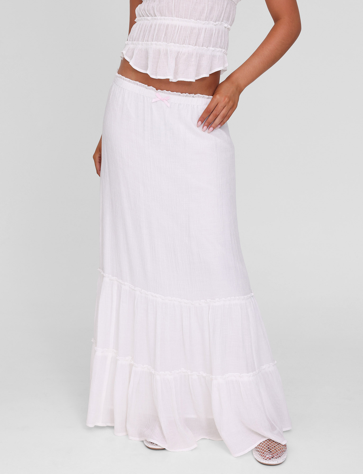 A person wears the TIGERMIST ASHLYNN MAXI SKIRT in white, featuring an elasticated waist, paired with a matching sleeveless ruffled top. The outfit is light and flowy; only the lower half of their face shows against a plain light gray background.