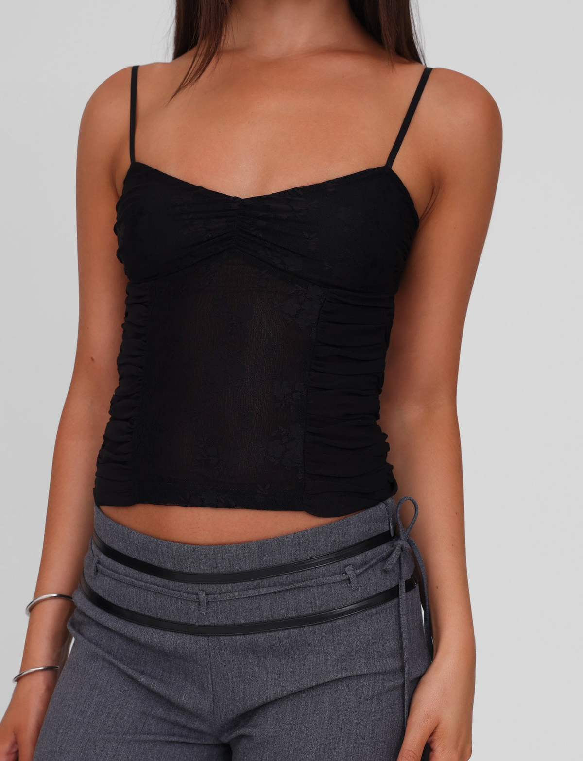 A person wears the TIGERMIST KALLIE TOP in black, a lace camisole with adjustable straps and ruched detailing, paired with gray pants featuring a side tie, standing against a plain background.