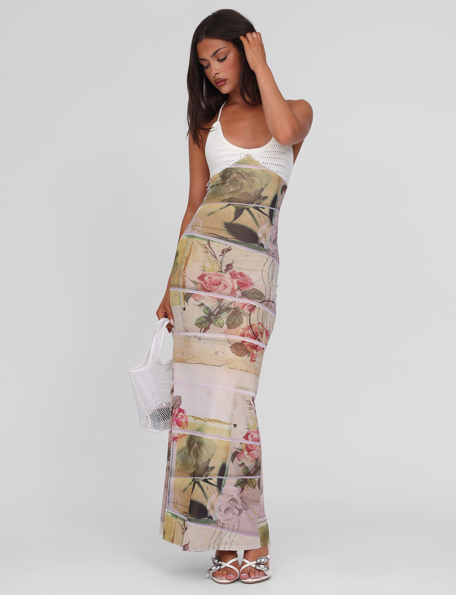 A woman wearing the TIGERMIST TILLEY MAXI DRESS - ROSE POSTCARD stands with a white mesh handbag and white sandals, touching her hair and posing against a plain light background.