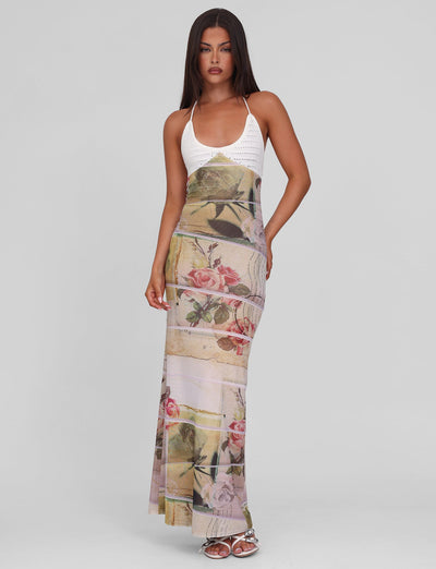 A woman with long dark hair and neutral makeup models the TIGERMIST TILLEY MAXI DRESS - ROSE POSTCARD, featuring crochet bust cups and a floral, abstract mesh skirt. She stands against a plain background wearing white sandals.