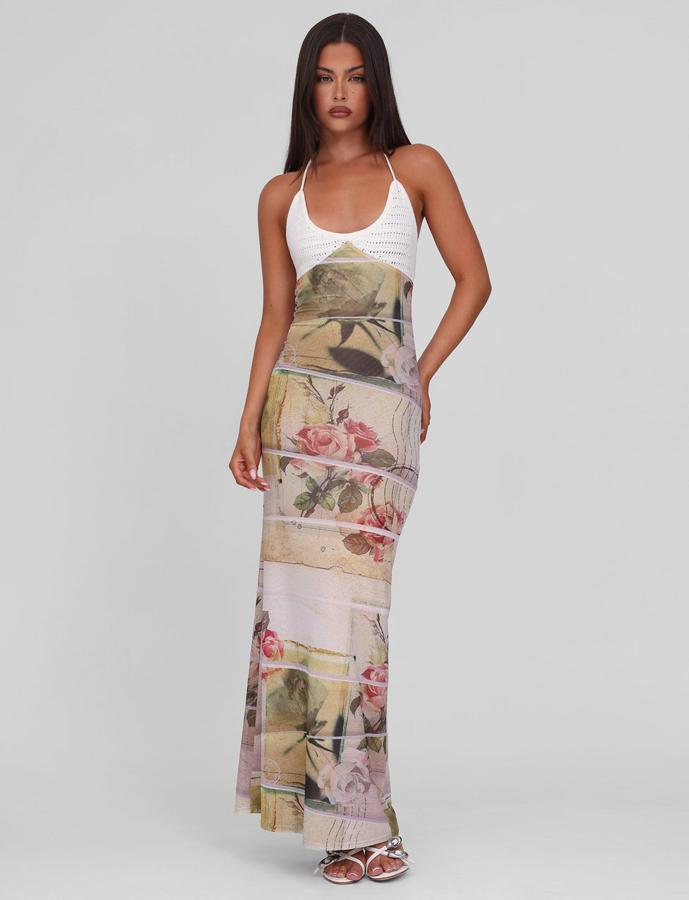 A woman with long dark hair and neutral makeup models the TIGERMIST TILLEY MAXI DRESS - ROSE POSTCARD, featuring crochet bust cups and a floral, abstract mesh skirt. She stands against a plain background wearing white sandals.