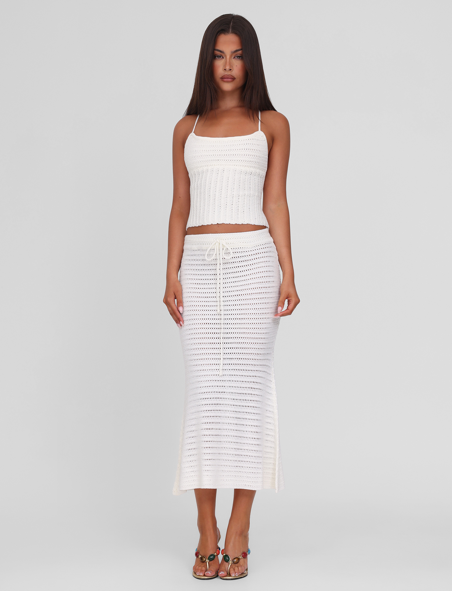 A woman with long, straight brunette hair wears the TIGERMIST EMELYN TOP in cream, a sleeveless crochet crop top with a lace-up back, paired with a matching ankle-length skirt and sandals against a plain white background.