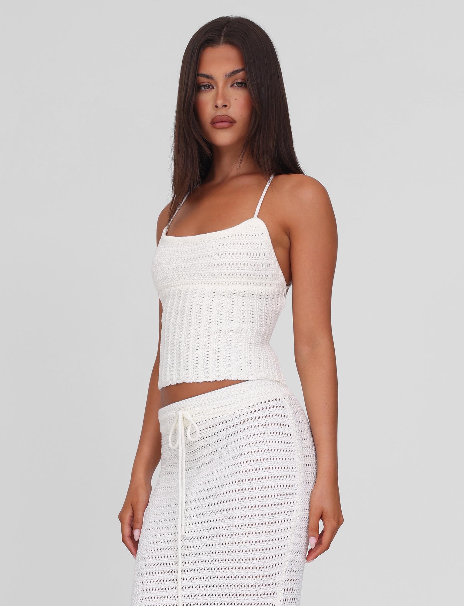 A woman with long dark hair wears the TIGERMIST EMELYN TOP in cream, featuring thin straps and a lace-up back, paired with a matching skirt, standing against a plain light gray background.