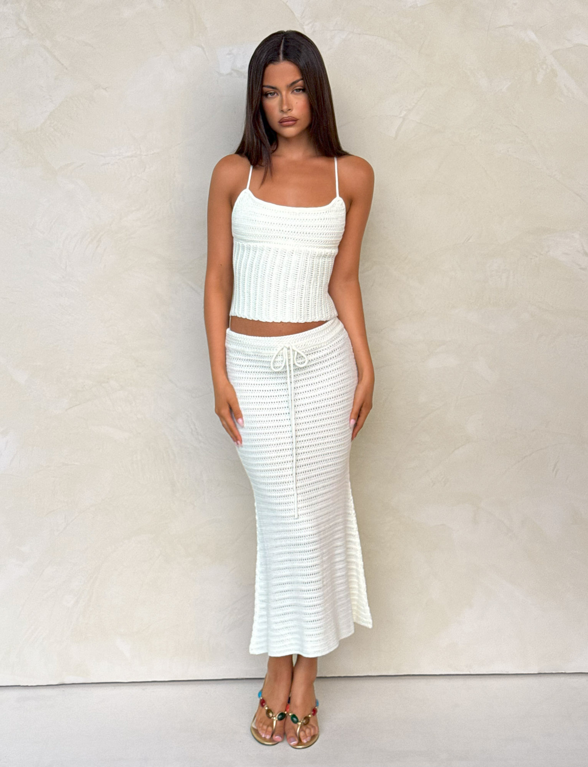 A woman with long dark hair stands against a textured, light wall wearing TIGERMIST’s EMELYN MIDI SKIRT - CREAM, styled with a white sleeveless knit top and open-toe sandals. The skirt features crochet fabric and an elasticated waist.