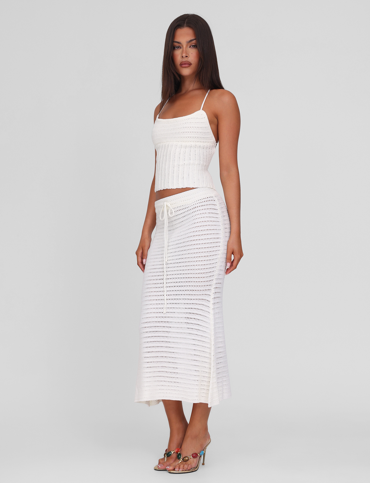 A woman with long brown hair wears the TIGERMIST EMELYN MIDI SKIRT in cream, featuring a crochet design and elasticated waist, paired with a white sleeveless ribbed knit top and heeled sandals against a plain background.